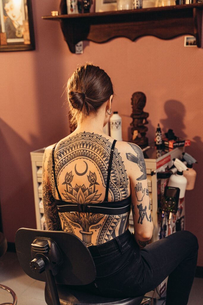 A woman with detailed body art designs sits in a tattoo studio, showcasing her intricate back tattoos.