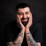 Portrait of a smiling man with tattoos leaning on a table in a studio. and Portrait of a smiling man with tattoos leaning on a table in a studio.